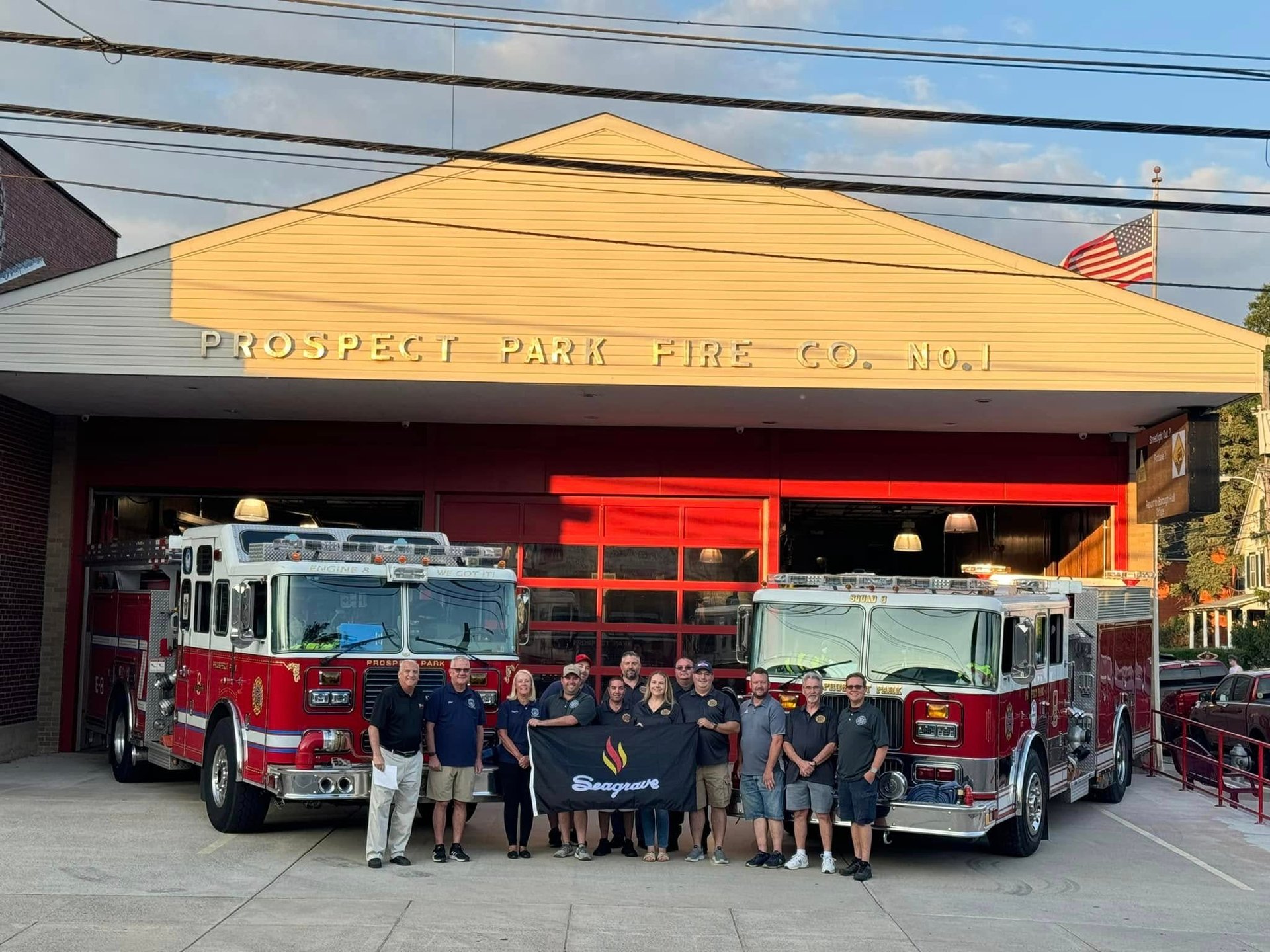 Group of firefighters standing in front of Prospect Park Fire Co. No. 1 station with red fire trucks