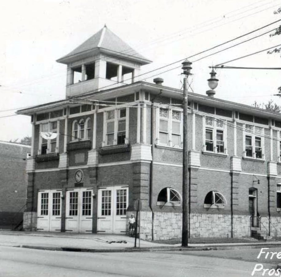Black and white photograph of a historic brick fire station with a tall tower and large garage doors on the ground floor