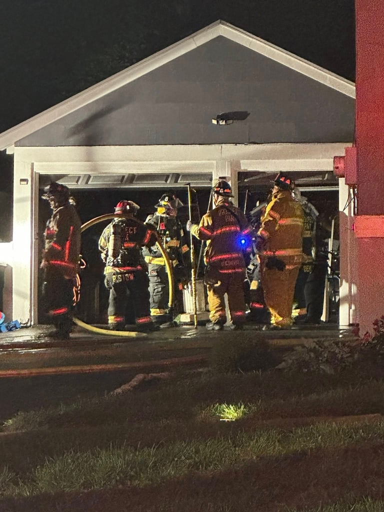 Firefighters in full gear gather inside an illuminated garage at night with equipment and hoses visible