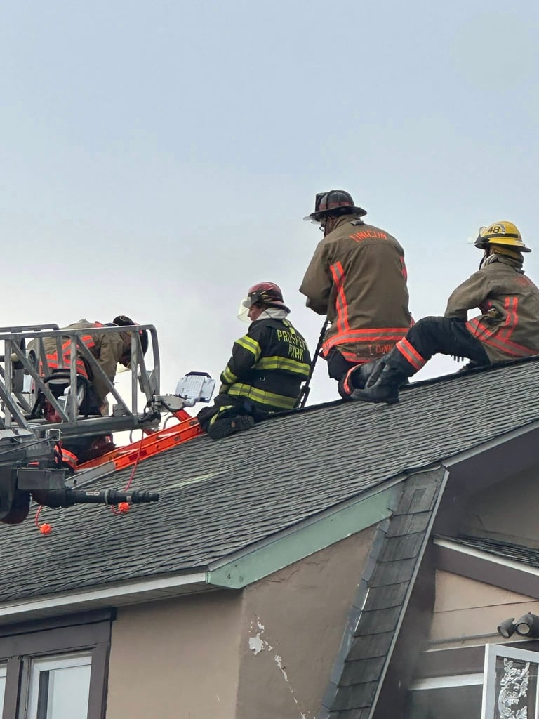 Firefighters in protective gear working on a residential roof with equipment and a fire truck visible