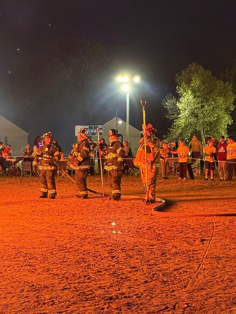 Firefighters in orange gear training on red dirt at night under bright floodlights with trees in background