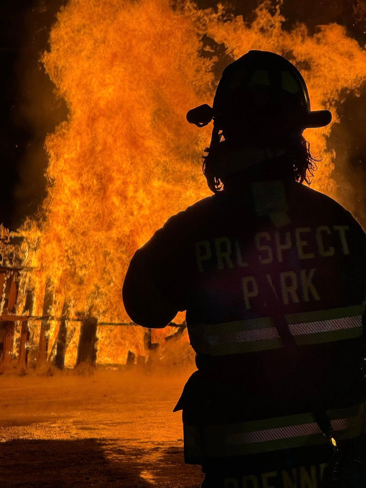 Firefighter in silhouette wearing helmet and reflective gear facing massive orange flames at night