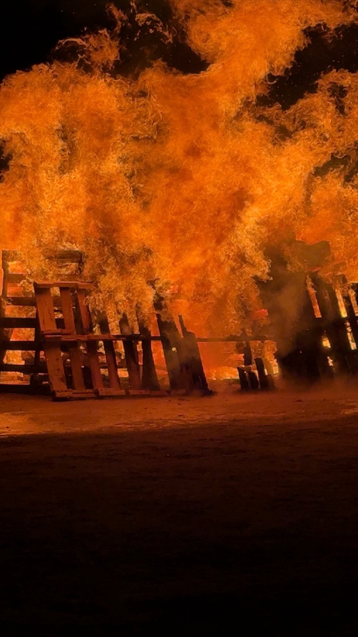 Massive flames engulf wooden structures and pallets in an intense fire against a dark night sky