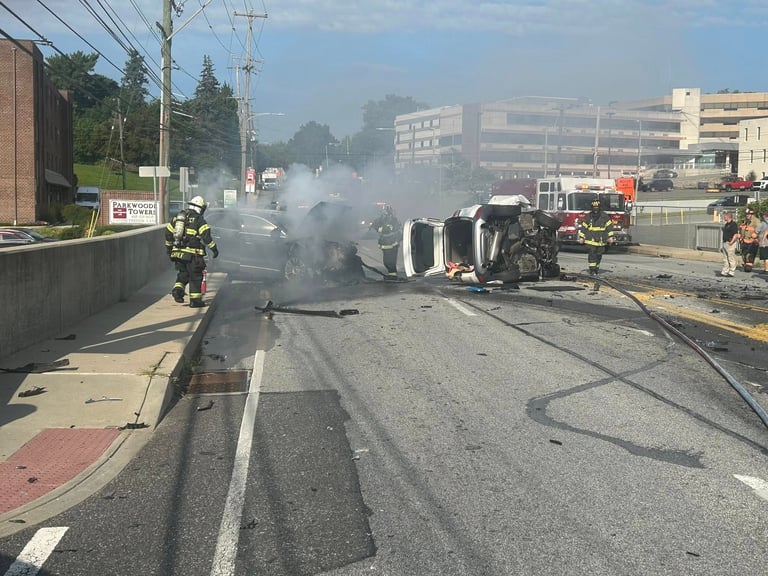 Overturned vehicle engulfed in smoke on city street with firefighters responding and emergency vehicles present