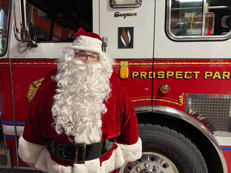 Santa Claus in full costume standing in front of a red fire truck labeled Prospect Park